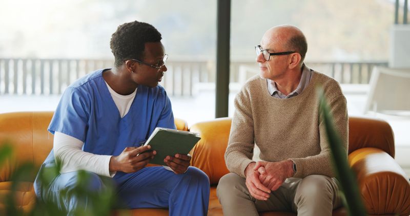 Tablet, conversation and nurse with mature man on sofa in home for diagnosis, discussion or treatment plan. Technology, consultation and medical caregiver with male patient in house for research.