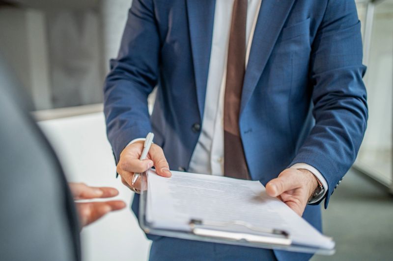 Business person in a suit holding a clipboard with a document and offering a pen, presenting paperwork for an agreement, deal, or contract in a professional setting