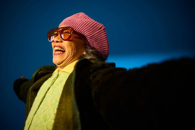 Senior woman wearing sunglasses and a beanie, laughing with arms wide open