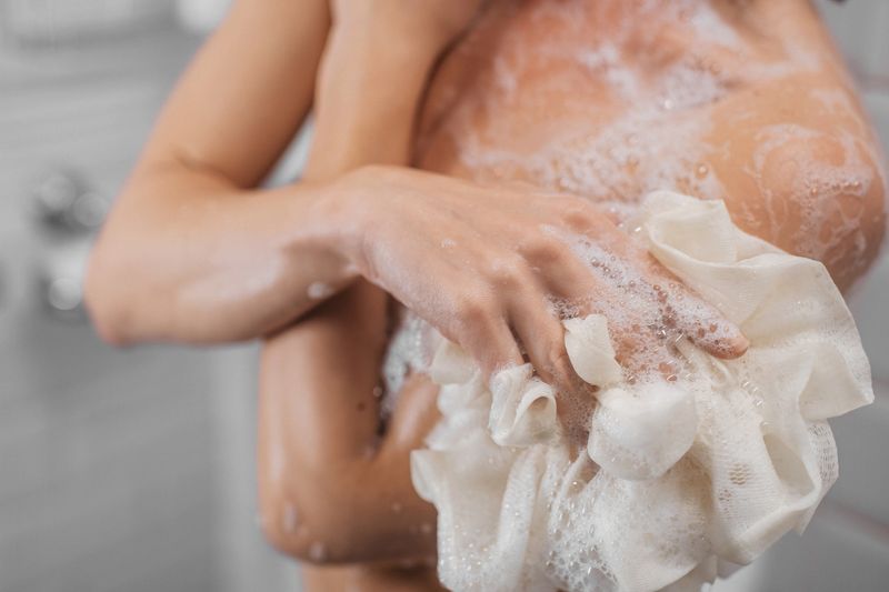 Female figure in shower, gently scrubbing skin with loofah, surrounded by bubbles and water droplets, emphasizing cleanliness and relaxation