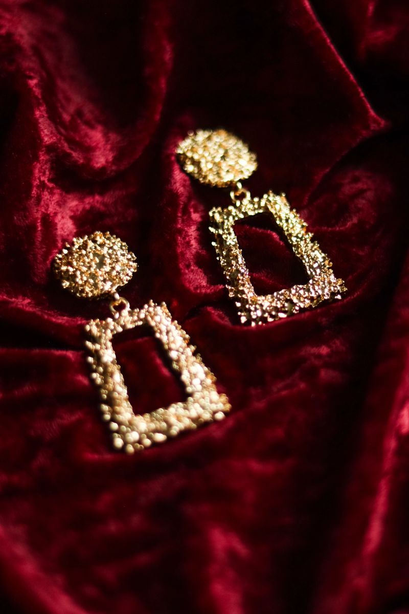 Two stylish gold colored metal drop earrings featuring a unique textured design, resting on a draped, soft deep red velvet surface