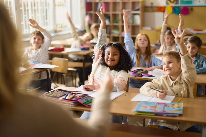 Large group of happy students raising their hands to answer the question teacher asked during a class at elementary school. Focus is on black girl.