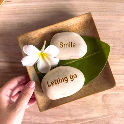 Two stones with words 'Smile' and 'Letting go' on a wooden tray with a flower and leaf.