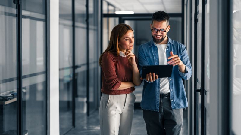 Two colleagues standing in a modern office hallway collaborate and discuss ideas using a tablet. The image portrays teamwork, technology usage, and a professional work environment.
