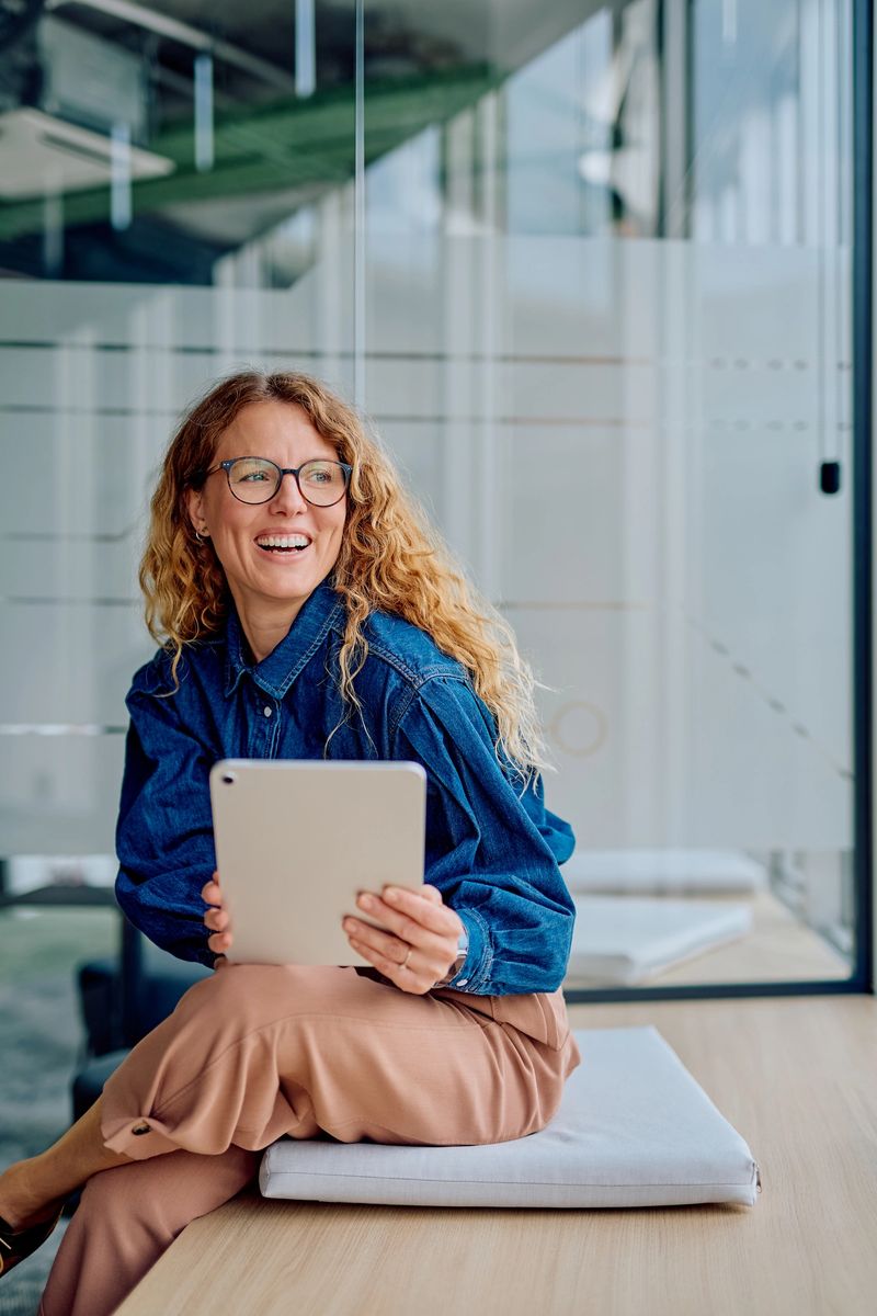 Happy professional woman using a tablet while sitting on a cushion in a modern office, enjoying successful mobile work