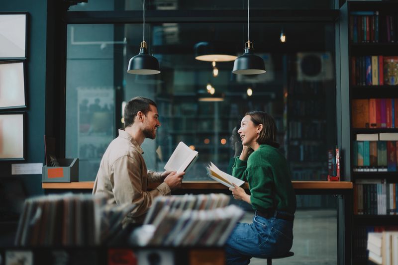 A relaxed adult couple sits at a counter in a library cafe, reading books and smiling as they chat, surrounded by shelves, warm pendant lights, and a calm, inviting atmosphere.