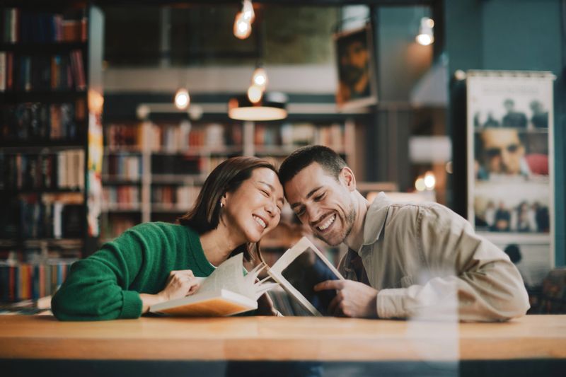 A joyful adult couple shares a warm moment in a cozy library cafe, reading together and smiling. Their close connection and happiness create a loving, inviting atmosphere.