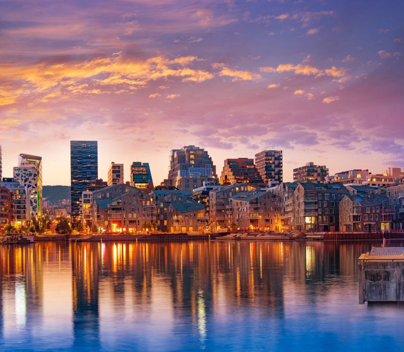 Modern waterfront apartments in Oslo Bispevika at summer sunset with warm lights reflecting on calm harbor water.