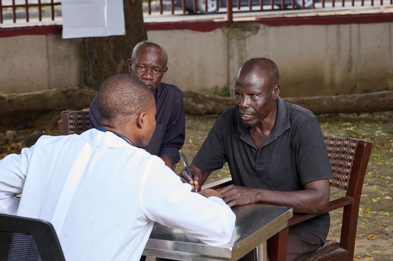 A young Nigerian doctor sits across a table from two older men during an outdoor medical consultation, listening and taking notes as one man speaks, with chairs and a community setting visible.