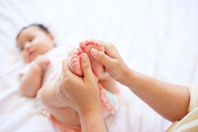 Close-up of a mother's hands tenderly holding and massaging the small feet of her baby girl lying on a white bedspread.