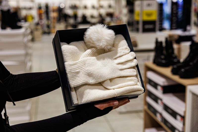 Person holding a boxed gift set containing a white knit hat with pom-pom and matching gloves, displayed in a clothing or shoe store