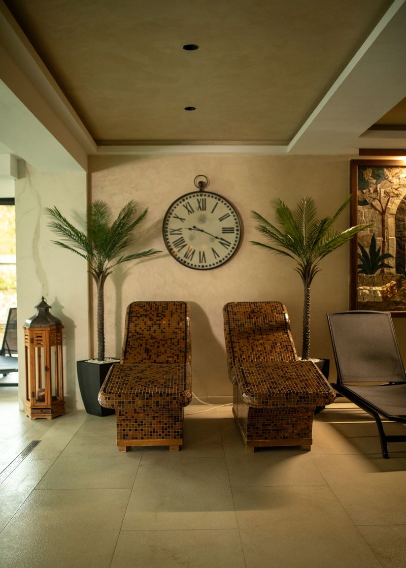 Interior of a tranquil wellness spa featuring mosaic tiled relaxation lounge chairs, tropical plants, and a large roman numeral clock under warm ceiling lights