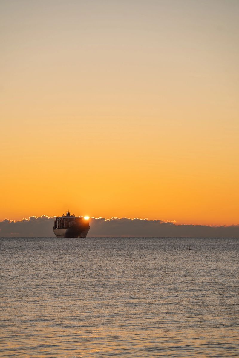 Sunrise behind vessel creates halo, Golden glow illuminates cargo ship with dramatic contrast, Morning sun casting striking silhouette over massive cargo vessel