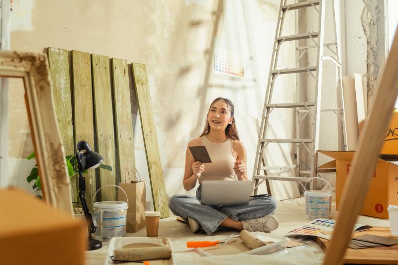 beautiful, pensive young woman (painter) sits on the floor of her new apartment, taking a break to drink coffee, daydreaming, and smiling while planning room color transformation with paint samples