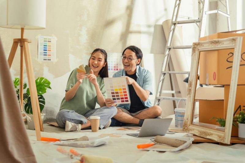 Collaborating with love and teamwork, a happy young couple takes a break from painting their apartment renovation to choose colors from a paint palette, aiming for a cozy, beautiful new home.