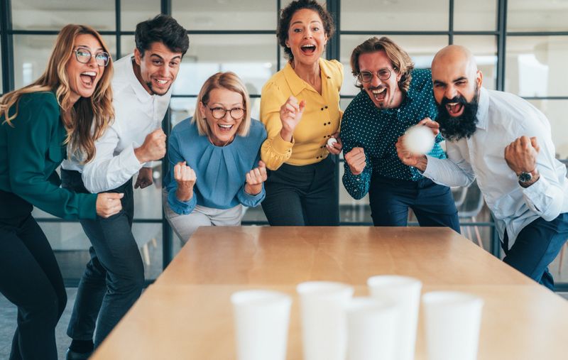 Group of cheerful creative business colleagues having fun while paying beer-pong in modern office