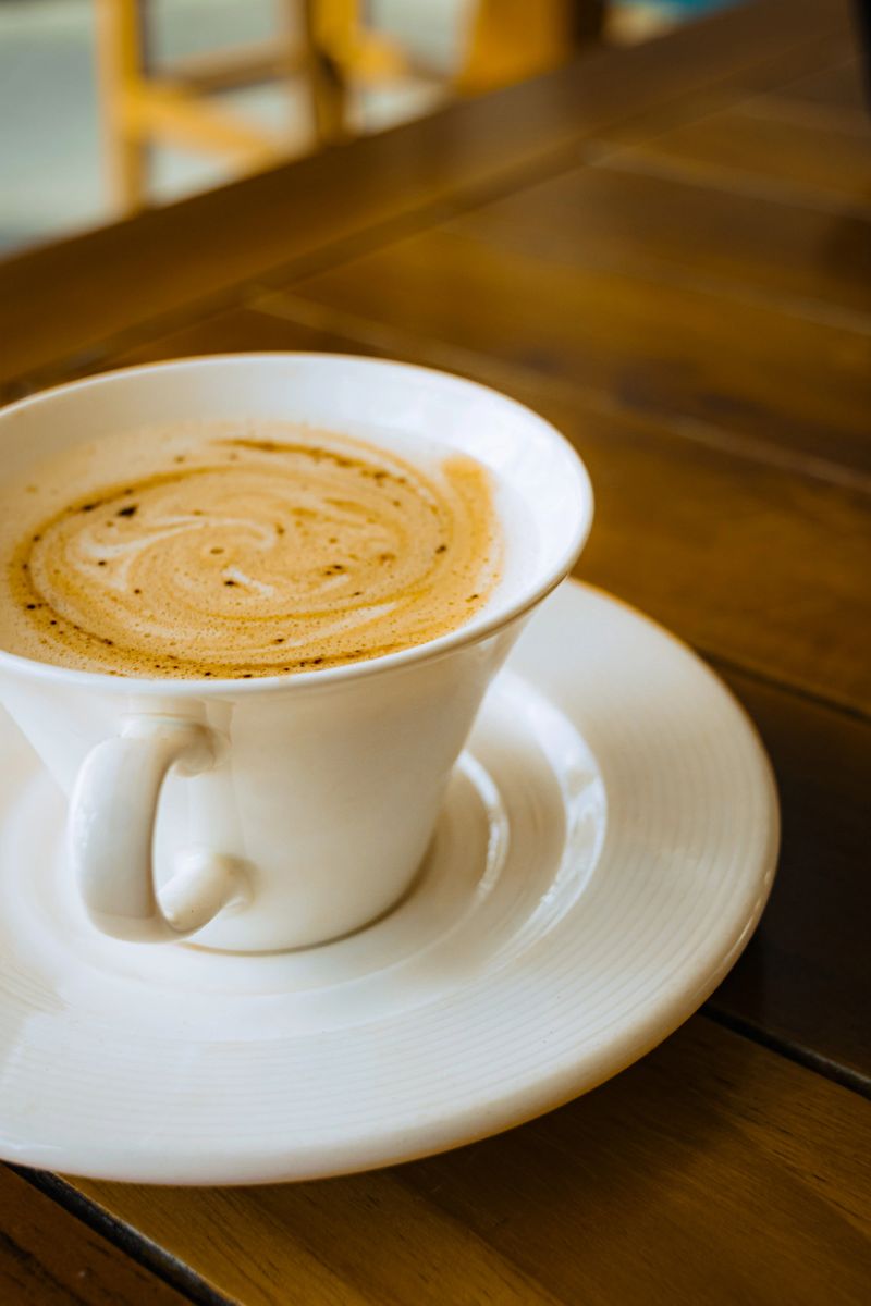 A ceramic coffee cup sits on a saucer atop a wooden table, showcasing intricate latte art in the foam. Soft lighting highlights the textures of the porcelain and wood.