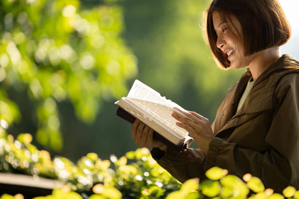 Woman reading a book outdoors with greenery around.