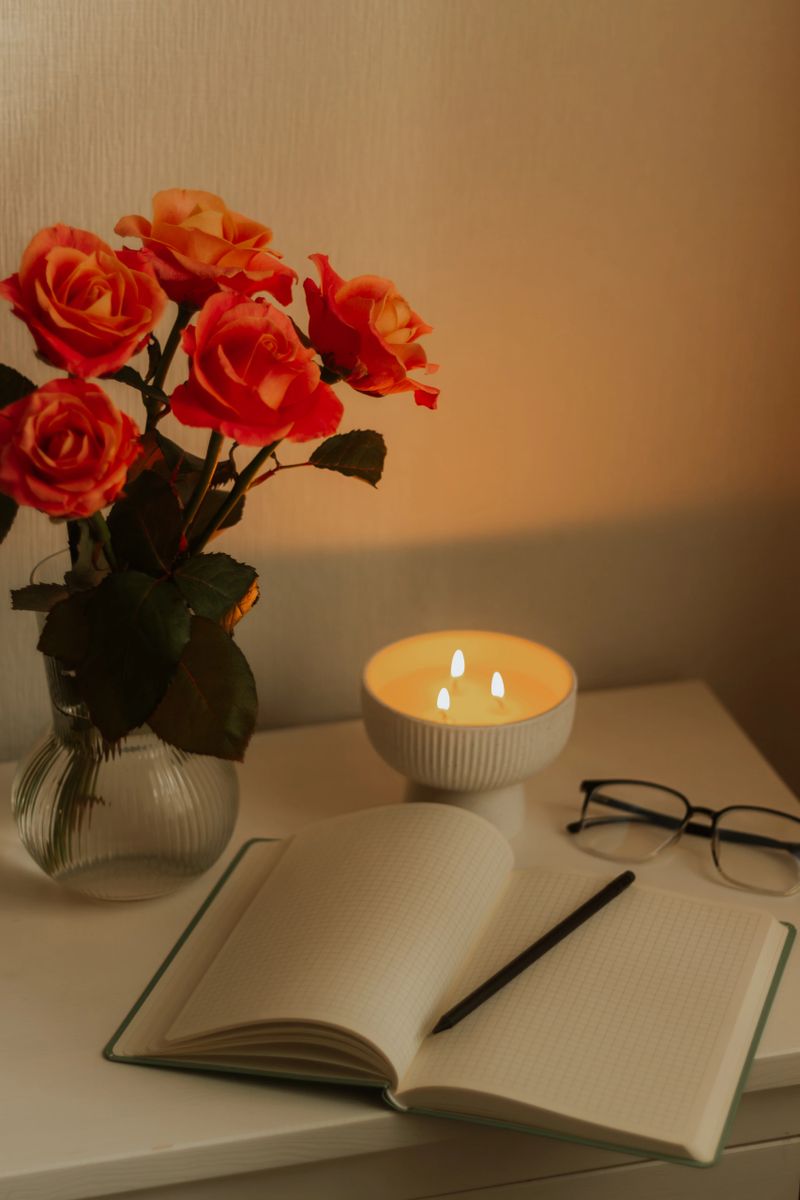 Warm cozy scene with glowing candles, roses in a vase, notebook and glasses on a white table. Soft evening light creates a calm atmosphere, highlighting peaceful home decor and a relaxing mood.