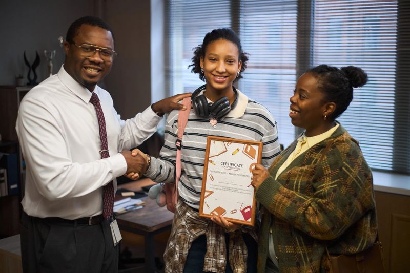 Middle aged Black man smiling and shaking hands with teenage Black girl holding certificate while middle aged Black woman standing beside them in office setting, celebrating achievement