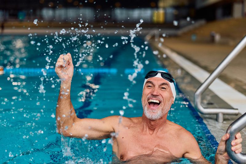 Smiling senior adult man with a swimming cap and goggles raising his arm, splashing water and celebrating his progress in an indoor pool, focusing on health and well-being