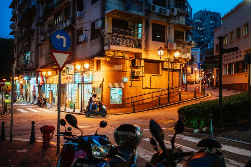 A lively Macau street at dusk with warm shop lights, scooters parked along the curb, and pedestrians in a busy urban corner. Curving road and vintage lamps create a welcoming city vibe.