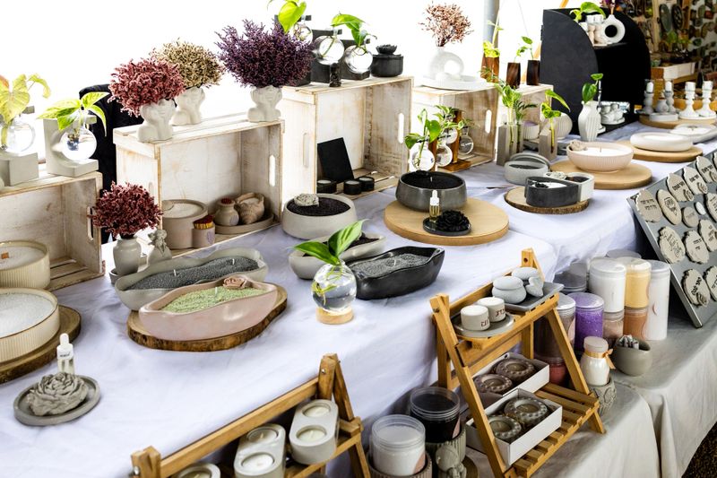 Elegant handmade candles in concrete and stone vessels arranged on a wooden table at a craft market. Minimalist natural decor with neutral tones, organic textures, and soft candlelight, reflecting slow living, hygge atmosphere, and modern handmade lifestyle design. Suitable for branding, interior decor concepts, and lifestyle backgrounds.