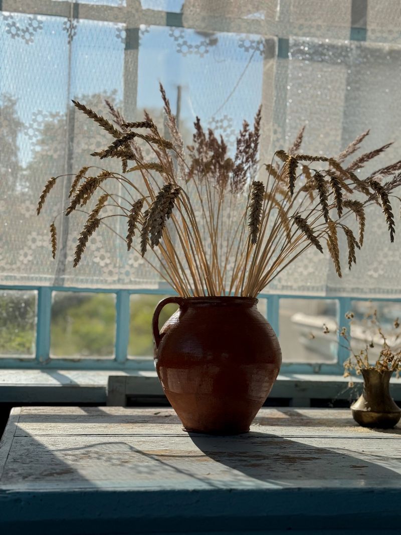 Rustic still life featuring dried plants in a ceramic pot on a wooden table near a window, lit by natural sunlight