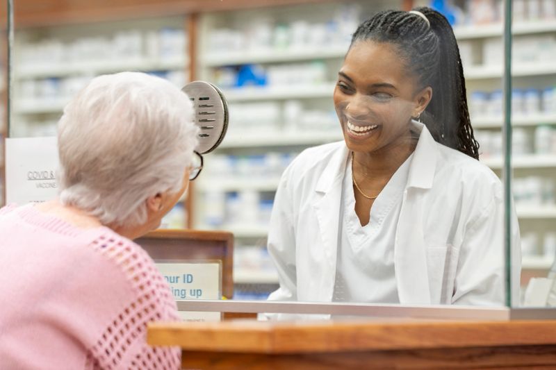 A smiling Black female pharmacist in a white coat chats with an elderly woman at a pharmacy counter, delivering warm, personable service in a modern drugstore setting today.