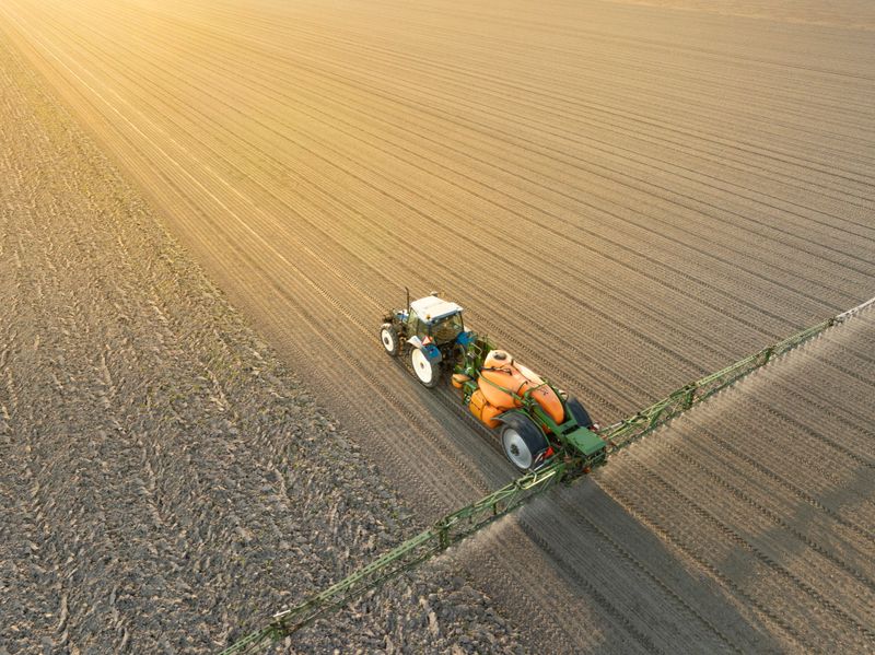 An aerial view captures a modern tractor pulling a large trailed crop sprayer across a vast agricultural field in Flevoland, Netherlands. The machinery is actively spraying herbicides, pesticides, or fertilizers, indicated by the fine mist emanating from the wide boom arms. The field, mostly bare earth with distinct furrows, stretches towards the horizon under a warm, golden sky, suggesting either early morning or late afternoon light during springtime. This image highlights contemporary farming practices, efficiency, and the use of advanced agricultural technology. It can represent themes of food production, environmental impact, sustainability debates, and rural development. Ideal for editorial content, agricultural industry publications, environmental reports, educational materials, and advertisements related to farming equipment, crop science, or land management.