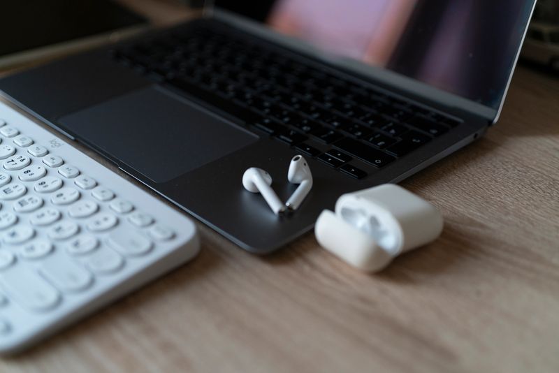 Earbuds sit beside a laptop and a wireless keyboard on a wooden table in a cozy setting.
