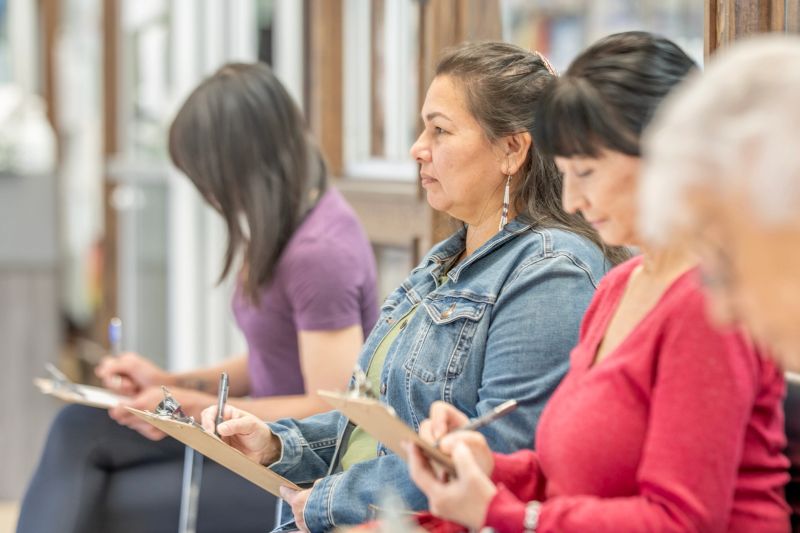A diverse group of women sits in a bright, indoor space, focused on clipboards and notes. The scene conveys learning, collaboration, and quiet concentration in a friendly workshop atmosphere.