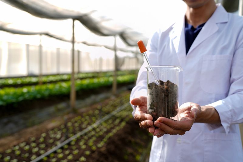 Asian man in lab coat and safety glasses putting soil sample into test tube doing research in greenhouse.