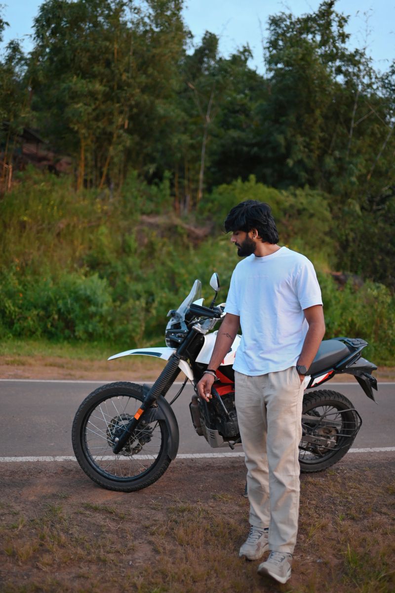 A young man stands beside his motorcycle on a quiet rural road surrounded by lush greenery. The scene captures a calm travel moment, highlighting road trips, freedom, adventure biking, and outdoor lifestyle in a natural countryside setting. Suitable for themes of travel, exploration, solo journeys, and modern lifestyle.