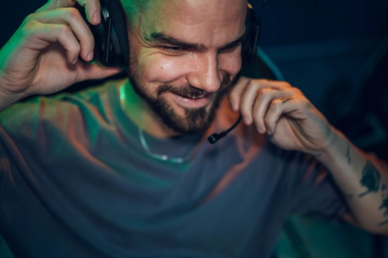Young adult male streamer with tattoos smiling and adjusting his gaming headset in a dark room with neon lights