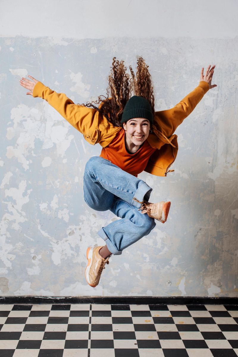 A young woman leaps mid-air in a bright orange jacket, jeans, and a green beanie; She exudes energy and joy in an urban, casual setting with a worn wall and checkered floor background