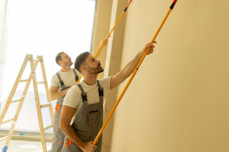Two men are painting a light-colored wall in a spacious room with large windows. They use long brushes and stand on a ladder, concentrating on their task as sunlight pours in.