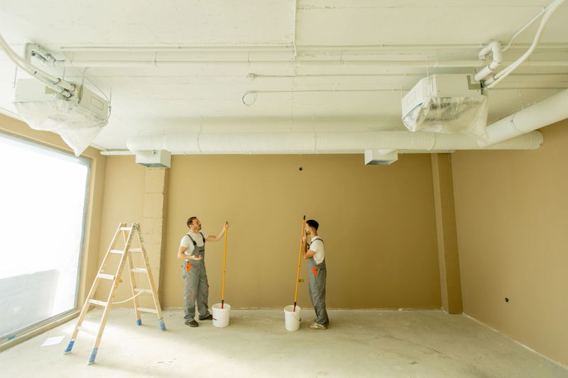 Two painters are focused on painting a wall beige in a well-lit room. They are using long rollers and standing on the floor next to a ladder and paint buckets, creating an inviting space.