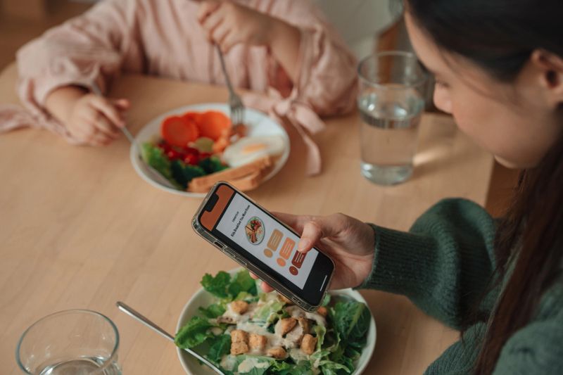 Asian woman using smartphone to plan weekly meals for her daughter with the help of a nutrition app while enjoying breakfast together at the table. As they share the meal, she balances technology and family time, ensuring her daughter's meals are nutritious and well-planned, while offering warmth and care to her little girl