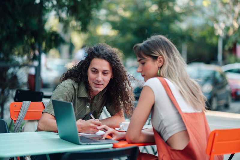 Young man and woman collaborating on project, looking at laptop screen and taking notes at an outdoor cafe