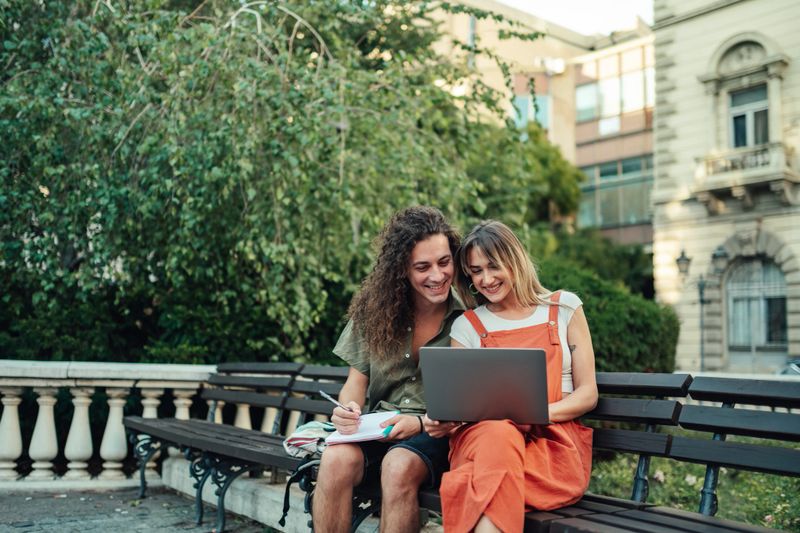 Young couple studying together on a park bench, smiling at a laptop while taking notes during a sunny campus day