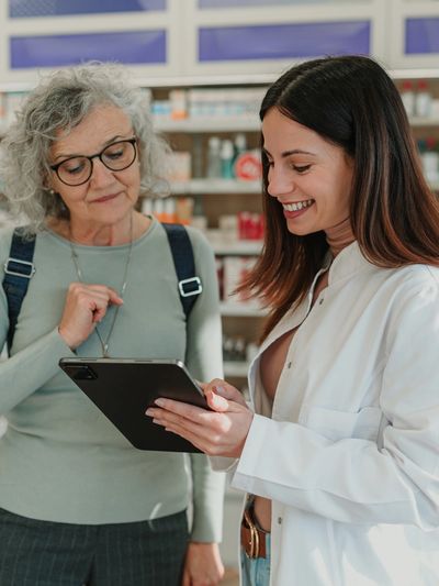 Pharmacist consulting with elderly woman using a tablet in a pharmacy.
