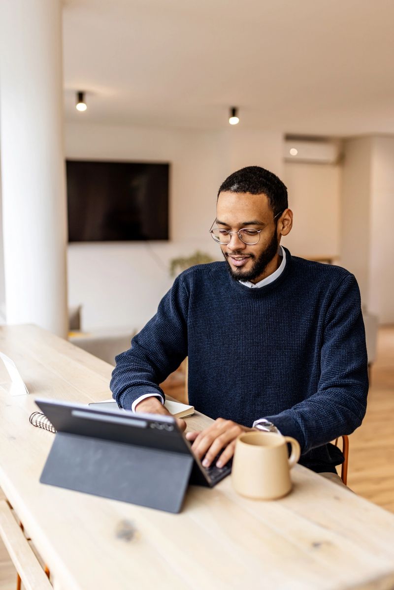 A man with glasses works on a tablet at a light wood desk in a bright, modern office. Calm, productive mood with a coffee mug and keyboard, suitable for business and startup visuals.