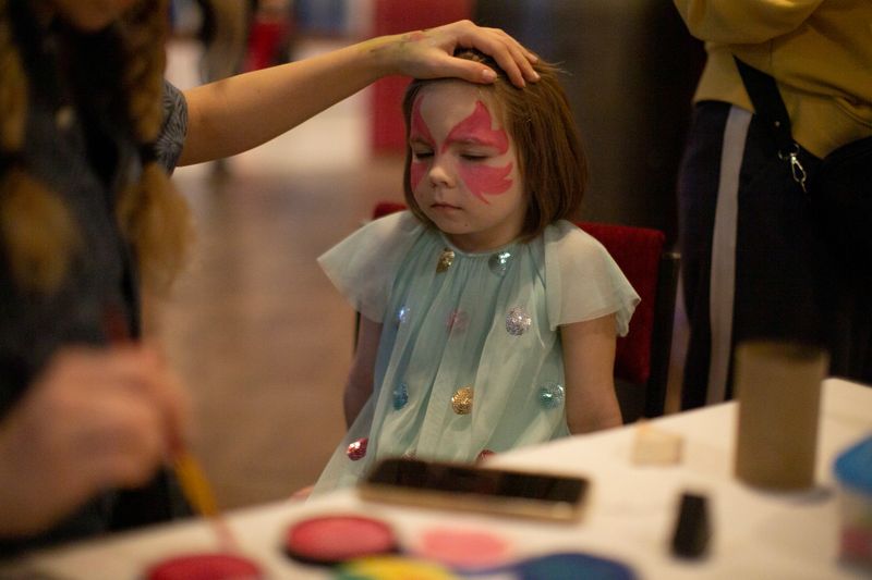 Young girl with butterfly face paint sits at craft table with palette and jars while artist finishes details, brushes and smartphone on table, adults