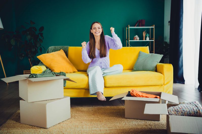 A cheerful woman sits on a vibrant yellow couch in a cozy living room with moving boxes, conveying a fresh start and warm home life in a stylish interior.