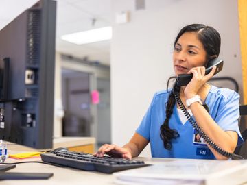 A nurse in blue scrubs talking on the phone while working at a computer.