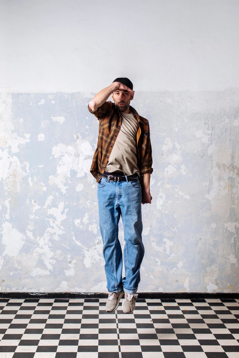 A casually dressed man in plaid shirt and jeans leaps in mid-air against a worn, textured wall and checkered floor, capturing energy, street style, and a playful urban vibe