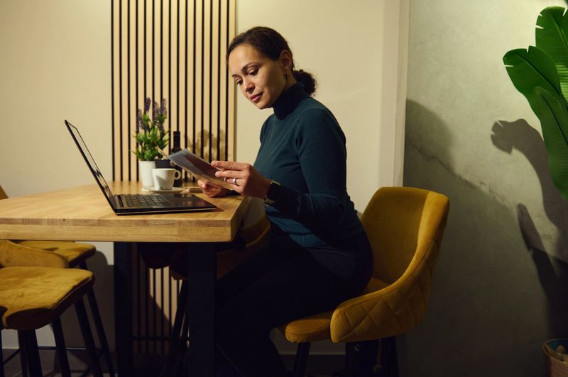 A professional Middle Eastern woman in a teal sweater works remotely at a minimalist cafe, using a laptop and tablet. Warm lighting, plants, and plush chairs create a chic, productive atmosphere.