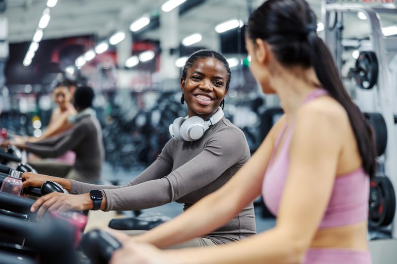 Two young women on stationary bikes in a gym, smiling and chatting as they cycle together, enjoying a friendly workout that promotes fitness, wellness, and fun companionship