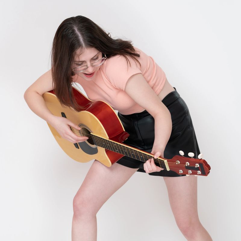 Emotional woman guitarist performs with playfulness and technical mastery, strumming guitar with authenticity. Playful woman wearing glasses, looking down at strings while playing musical instrument